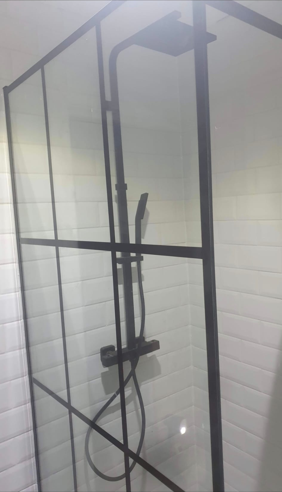 Walk-in shower with black-framed glass screen, black thermostatic riser rail and handheld head against white subway tiling, electric shower installation, London.